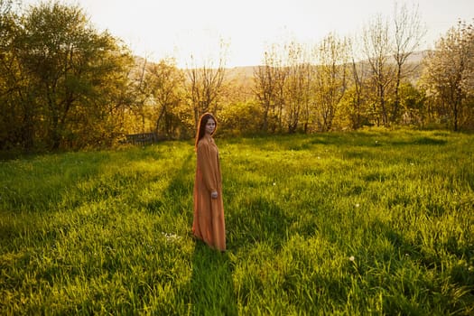 an attractive, slender, red-haired woman stands in a wide green field during sunset in a long orange dress enjoying unity with nature and relaxation. High quality photo