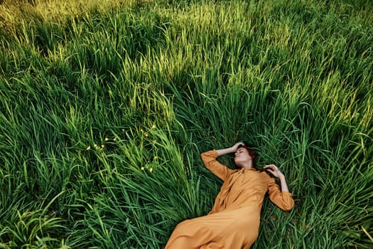 a sweet, calm woman in an orange dress lies in a green field with her hands under her head enjoying silence and peace. Horizontal photo taken from above. High quality photo