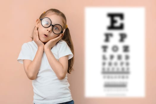 Surprised girl 5 - 7 years old in round glasses on the background of a blurred snellen table for checking visual acuity.