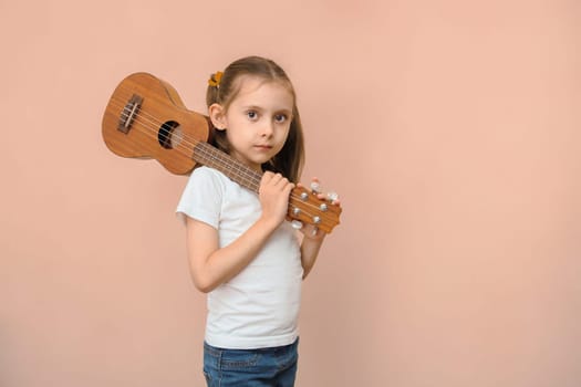 Caucasian girl in a white T-shirt with a ukulele on his shoulder looks at the camera on a pink background.