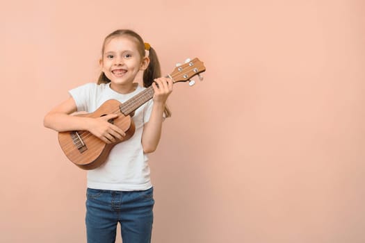 Happy caucasian smiling girl 5 -7 years old in a white t-shirt with a ukulele on a pink background, copy space.