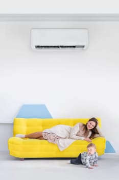 Young caucasian woman with child lies on a yellow sofa in bright interior with split air conditioner.