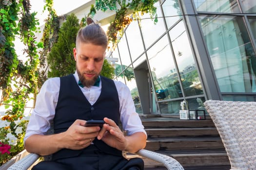 Young hipster in a white shirt, black vest looks into a smartphone while sitting in a penthouse.