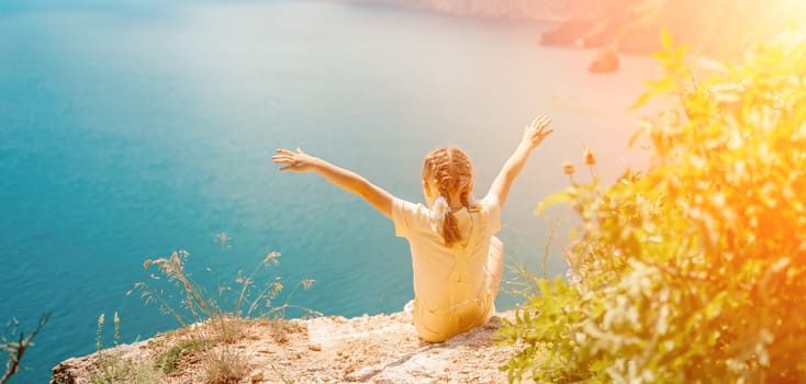 Happy girl perched atop a high rock above the sea, wearing a yellow jumpsuit and braided hair, signifying the concept of summer vacation at the beach