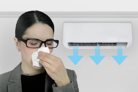 A woman with a tissue to her nose is having a cold and feeling unwell due to the air conditioner in the office.