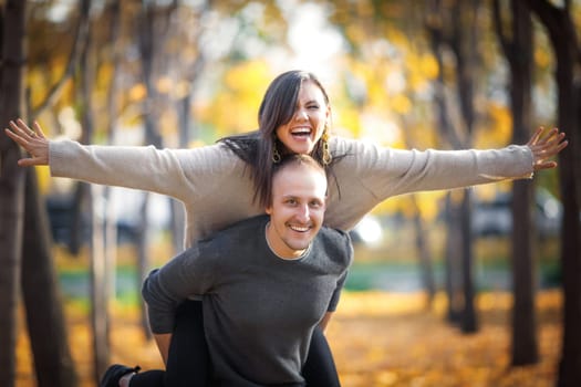 Happy caucasian man carrying his asian girlfriend on back in autumn outdoor.