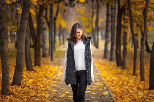 Young mixed race woman in autumn park along the alley with fallen autumn leaves.