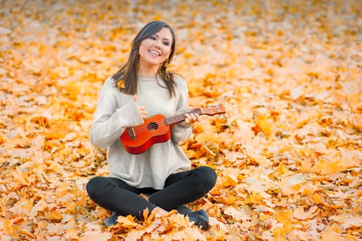 Young happy mixed race woman playing ukulele in autumn park.