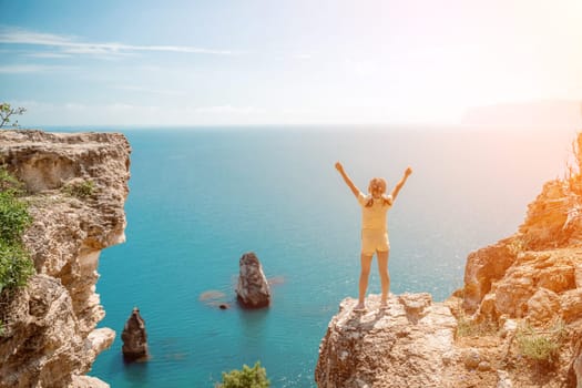 Happy girl stands on a rock high above the sea, wearing a yellow jumpsuit and sporting braided hair, depicting the idea of a summer vacation by the sea