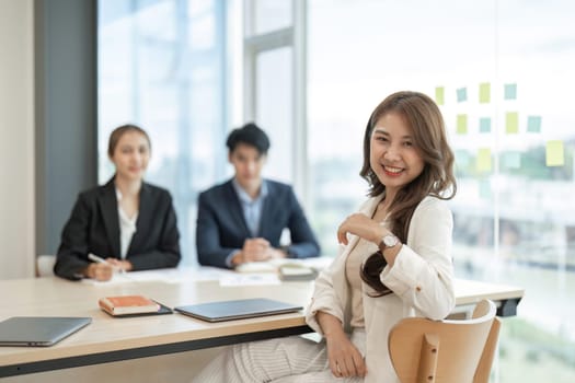 portrait of young asian business woman standing in office arms crossed with coworkers colleague talking in background.