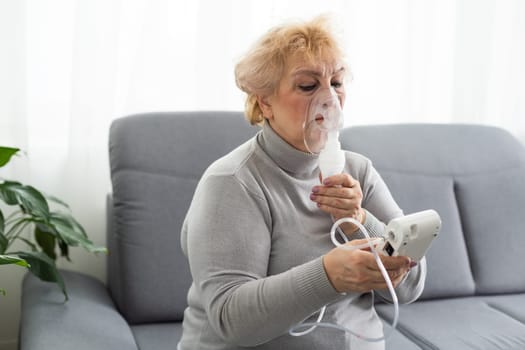 Senior woman using a nebulizer makes inhalation at home and looks at the camera.