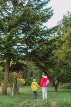 Beautiful family -Mom with small son on a walk in autumn sunny nature. Happy couple holding hands. Back view. Young mother with her little baby boy having fun in the autumn park