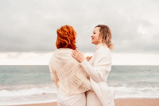 Women sea walk friendship spring. Two girlfriends, redhead and blonde, middle-aged walk along the sandy beach of the sea, dressed in white clothes. Against the backdrop of a cloudy sky and the winter sea. Weekend concept