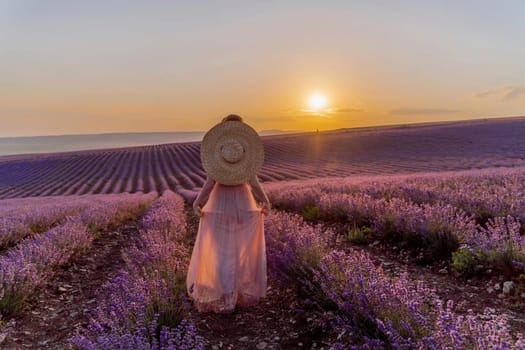 Woman lavender field sunset. Romantic woman walks through the lavender fields. illuminated by sunset sunlight. She is wearing a pink dress with a hat