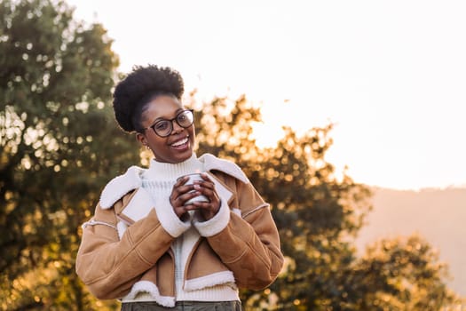 young african woman smiling happy in nature with a mug of hot drink in hand, concept of morning bliss and wilderness lifestyle, copy space for text