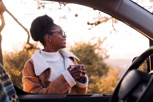 young african woman traveling in a camper van smiling happy in nature with a mug of hot drink in hand, concept of morning bliss and wilderness lifestyle