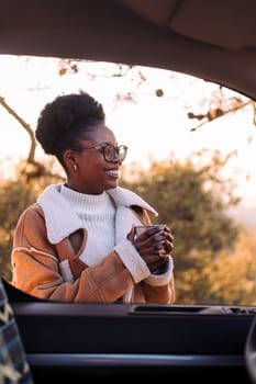 young african woman traveling in a camper van smiling happy in nature with a mug of hot drink in hand, concept of morning bliss and wilderness lifestyle
