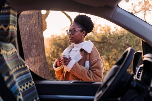 young african woman in a road trip in camper van embraces the outdoors finding peace with tea in the morning, concept of nature's warmth and wilderness lifestyle