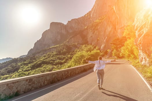 woman running mountain. Happy woman runs along an asphalt road in the middle of a mountainous area. She is dressed in jeans and a white shirt, her hair is braided. A traveler in the mountains rejoices in nature