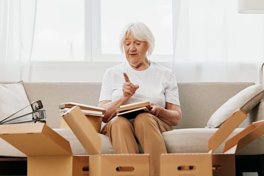 elderly woman sits on a sofa at home with boxes. collecting things with memories albums with photos and photo frames moving to a new place cleaning things and a happy smile. Lifestyle retirement. High quality photo