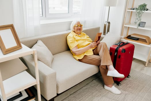 Happy senior woman with passport and travel ticket packed a red suitcase, vacation and health care. Smiling old woman joyfully sitting on the sofa before the trip raised her hands up in joy. High quality photo