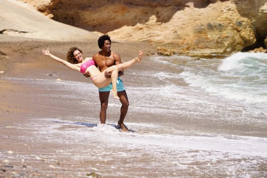Full length of young African American male in swimwear raising female with outstretched arm while enjoying summer holidays on beach