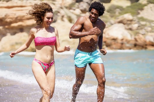 Smiling young African American male running with female companion in splashing water and looking away while enjoying fun together on seashore during sunny day