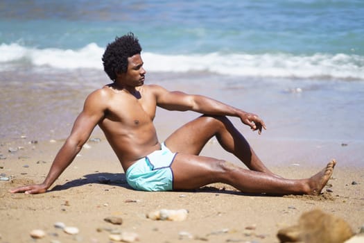 Full body side view of African American male relaxing on sandy beach with arm on knee while looking away, and leaning on hand near waving sea