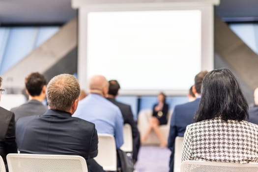 Business and entrepreneurship symposium. Female speaker giving a talk at business meeting. Audience in conference hall. Rear view of unrecognized participant in audience.