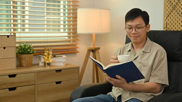 Calm asian man wearing glasses sitting on sofa and reading book. Leisure and people concept.