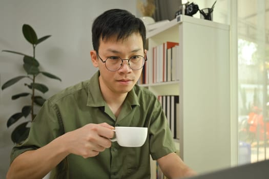 Concentrated asian man freelancer wearing glasses drinking coffee and reading email on laptop computer.
