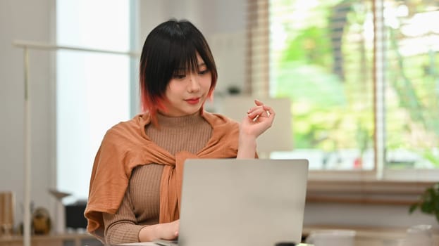 Charming young asian woman in stylish clothes working on laptop computer at modern home office.