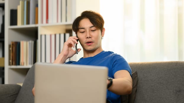 Smiling handsome man having phone conversation, using laptop while sitting on couch in living room.