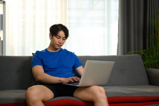Image of handsome asian man wearing casual clothes using laptop on couch at home.