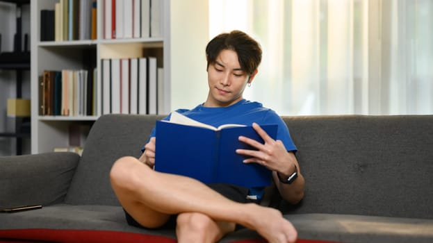 Smiling asian man relaxing on couch and reading book. Leisure and people concept.