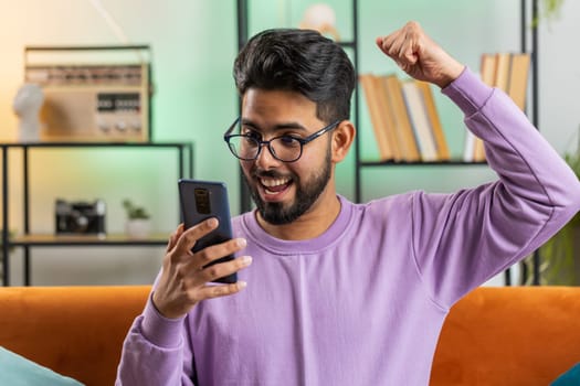 Happy indian man use mobile smartphone typing browsing say Wow yes found out great big win good news celebrate lottery jackpot doing winner gesture. Hindu young guy at home in room sitting on sofa
