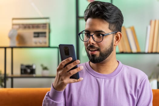 Indian man sitting on sofa uses mobile phone smiles at home living room apartment. Hindu young guy texting share messages content on smartphone social media applications online, watching relax movie