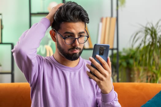 Sad displeased indian man use smartphone typing browsing, loses becoming surprised sudden lottery results, bad news, fortune loss, fail, deadline, virus. Young hindu guy at home living room on couch