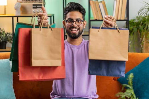 Young indian man happy shopaholic consumer came back home after shopping sale with bags. Portrait of hindu guy satisfied received parcels purchase from online order at home apartment room on sofa
