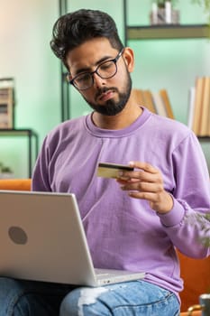 Young indian man using credit bank card and laptop computer while transferring money, purchases online shopping, order food delivery. Hindu guy at home apartment living room on couch. Vertical