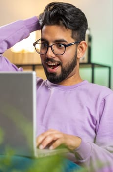 Surprised indian man guy using laptop computer, receive mail good news message, shocked by sudden victory celebrate lottery jackpot win purchases online shopping at home apartment on sofa. Lifestyles