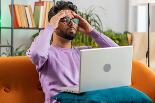 Tired freelancer indian man use laptop, suffering from headache problem tension and migraine, stress at home room. Hindu guy works on notebook, sends messages, makes online purchases, watching movies