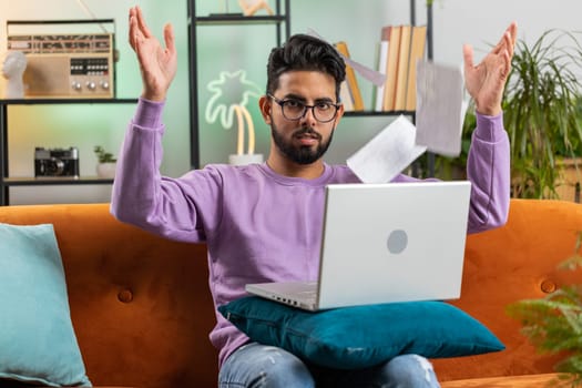 Stressed indian man at home room looking at unpaid bank debt bills, doing paperwork, planning budget, calculate finances mortgage payments. Hindu guy throws paper bills sitting on couch. Lifestyles