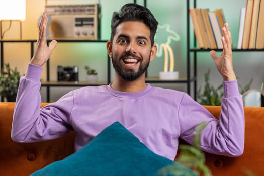 Happy indian man shouting, celebrating success, winning, goal achievement good news, lottery luck, victory, watching TV championship at modern home apartment. Hindu guy in living room sitting on couch