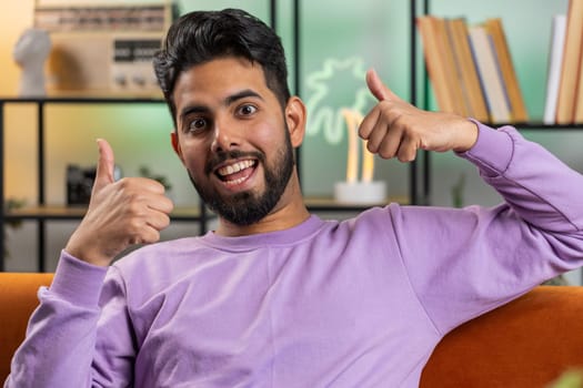 Like. Happy excited indian man looking approvingly at camera showing thumbs up, like sign positive something, good great news, positive feedback. Young hindu guy sitting on couch at home living room