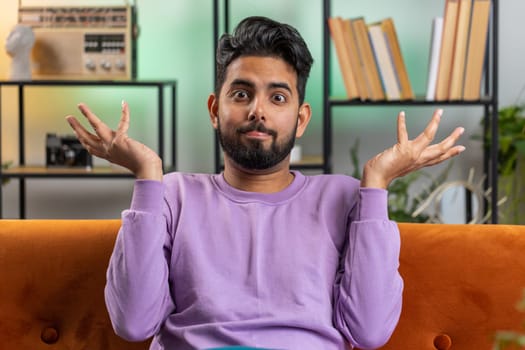 I dont know what to say. Confused indian handsome man feeling embarrassed about ambiguous question, having doubts, no idea, being clueless and uncertain at home. Hindu guy sits on sofa in living room