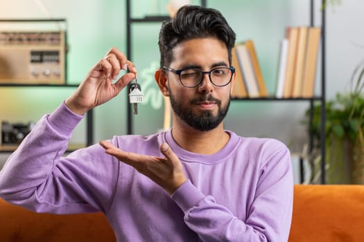 Happy young indian man real estate agent lifting hand showing the keys of new home house apartment, buying or renting property, mortgage loan. Portrait of hindu guy at home apartment room on sofa