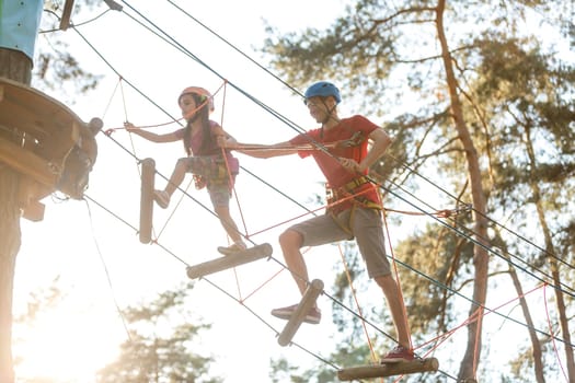 Young father helps his daughter on the obstacle in the rope park.