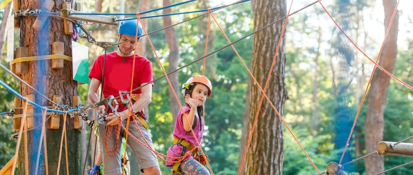 Young father teaching her daughter to climb on the rock climbing wall. Little girl preschooler wearing safety harness having fun time in adventure rope park. Happy family concept.