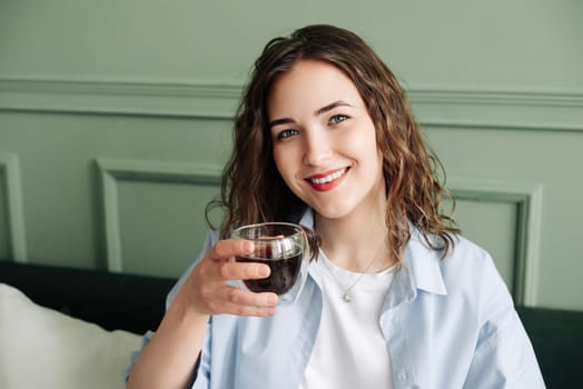 Happy young woman housewife enjoying a cozy break with hot tea on the sofa at home. Smiling lady relaxing with a warm drink and looking at the camera in a close-up indoor portrait